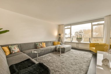 Gri koltukları ve sarı sandalyeleri olan büyük bir pencerenin önünde sokağa bakan bir oturma odası.