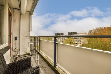 Güneşli bir günde arka planda birkaç sandalye ve ağaç olan bir balkon, bir apartman penceresinden görüldüğü gibi.