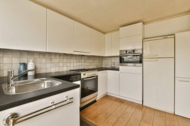 Bu fotoğrafta tezgahların üzerinde beyaz dolaplar ve siyah tezgahlar olan bir mutfak içeriden çekilmiş.