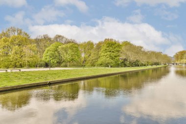 Parkın ortasında bir nehir, her iki tarafında ağaçlar ve durgun suya yansıyan mavi gökyüzü.