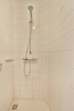 Duş başlıklı beyaz fayanslı bir banyo ve duvardaki parmaklıklar yerdeki seramik fayanslardan yapılmıştır.