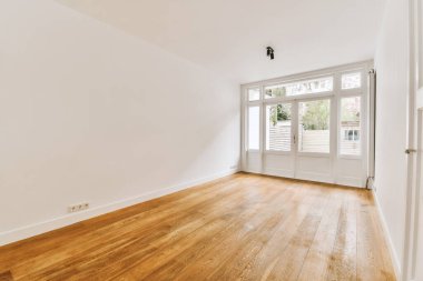 Ahşap döşemesi ve duvarlarında beyaz boya olan boş bir oda. Başka bir odaya açılan bir kapı var.