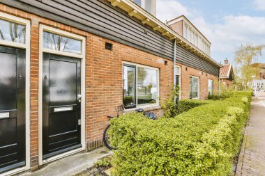 Binanın yan tarafında iki siyah kapısı ve çalıları olan tuğladan bir bina önünde park edilmiş bir bisiklet var.