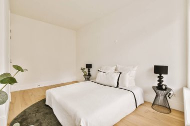 Beyaz duvarları ve parke döşemeleri olan bir yatak odası. Pencereden gelen ışıkla aydınlanıyor.