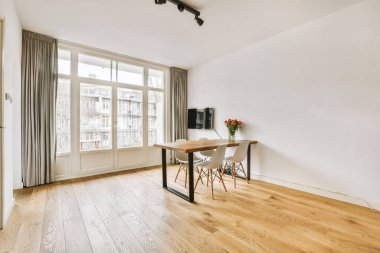 Oturma odasında bir yemek masası ve sandalyeler. Ahşap zeminler, beyaz duvarlar ve sokağa bakan büyük pencereler.