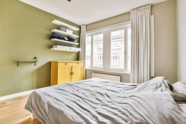 Yeşil duvarlı ve penceresinde beyaz süslemeler olan bir yatak odası. Sarı göğüs dolabının önünde.