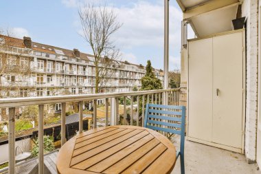 Arka planda binalar olan bir balkonda bir masa ve sandalyeler. Bir apartmanın penceresinden sokağa bakılıyor.