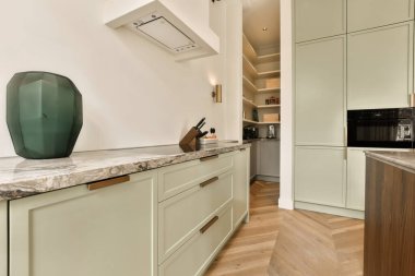 Mermer tezgahlı modern bir mutfak ve beyaz dolaplar pencereden gelen ışıkla aydınlanır.