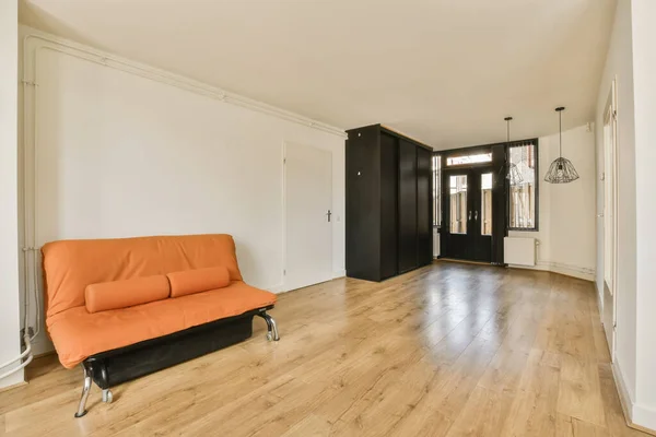 a living room with hardwood flooring and an orange couch in the center of the room is a white wall
