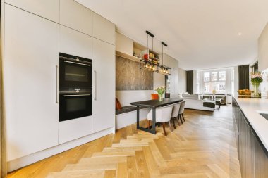 Odanın ortasında ahşap döşemeli ve beyaz dolaplı modern bir mutfak bir fırın, yemek masası ve sandalyelerle dolu.