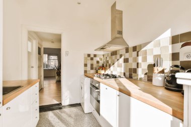 Resmin ortasında beyaz dolaplar ve ahşap tezgahlı bir mutfak başka bir odaya açılan açık bir kapıdır.