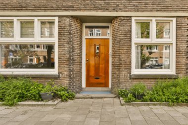 Güneşli bir günde aşağıdaki sokaktan görüldüğü gibi ön bahçesinde iki pencere ve bitkiler olan tuğladan bir ev.