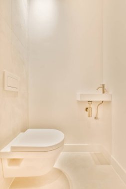 Beyaz duvarları ve bej döşemeleri olan modern bir banyo. Duvarda da bir tuvalet var. Işıklandırmayla aydınlatılıyor.