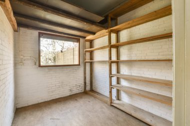 Beyaz tuğlalı duvarları ve duvarında ahşap rafları olan boş bir oda görünüyor. Köşede bir pencere var.