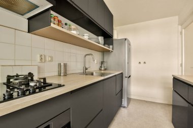 Arkasında siyah dolaplar ve beyaz fayanslar olan bir mutfak başka bir odaya açılan açık bir kapıdır.