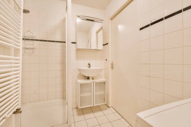 Küvetin etrafında beyaz fayanslar ve siyah süslemeler olan küçük bir banyo, lavabo ve duvarda ışıldayan bir raf.