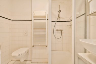 Beyaz fayanslı duvarları ve zemini olan küçük bir banyo. Duş kabininin arasındaki köşede de tuvalet var.