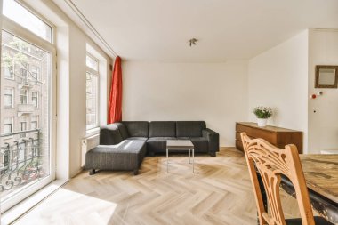 Büyük bir pencerenin önünde ahşap döşemeli ve siyah kanepeli bir oturma odası var.