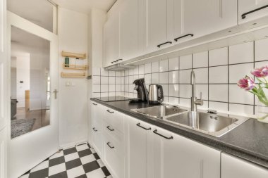 Beyaz bir mutfak, yerde siyah beyaz kareli fayanslar ve odanın ortasında bir lavabo.