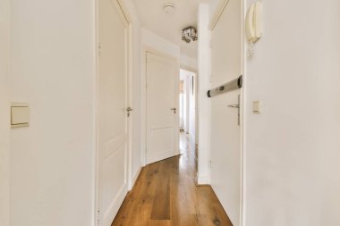 Beyaz duvarları ve ahşap döşemeleri olan boş bir koridor pencerelerden gelen doğal ışıkla aydınlanır.