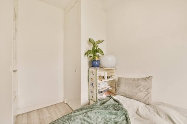 Beyaz duvarlı ve tahta döşemeli bir yatak odasında küçük bir yatak odası ve yanında da bir bitki var.