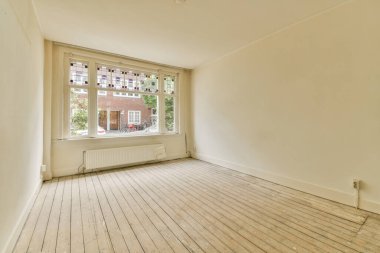 Büyük bir pencerenin önünde beyaz duvarları ve ahşap döşemeleri olan boş bir oda sokağa bakıyor.