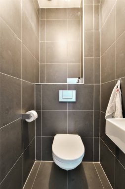 Duvarında gri fayans duvarları ve beyaz tuvaleti olan küçük bir banyo, tuvaletin üstündeki ışıklarla aydınlatılıyor.