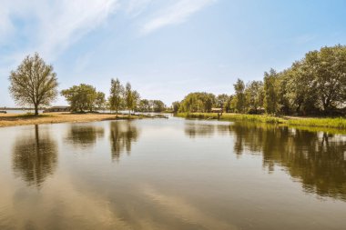 Arka planda ağaçlar ve üzerinde mavi gökyüzü olan bir nehir, suların ötesinden görüldüğü gibi.