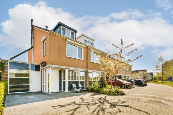 stock image a house in the middle of a residential area with cars parked on the driveway and trees growing along the road