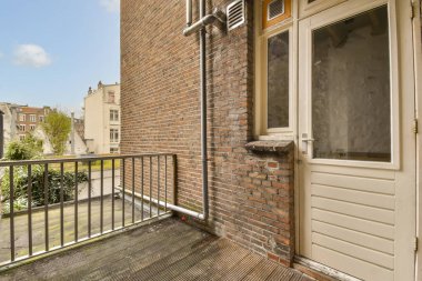 Arka planda tuğla bina ve kapısı açık bir balkon var. Dışarıdan sokağa bakıyor.