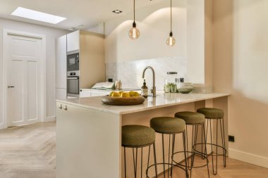 Üç taburesi ve tezgahın önündeki adada bir kase meyvesi olan bir mutfak beyazdır.