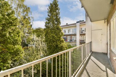 Önünde ağaçlar ve çalılar olan bir balkon. Pencereli bir alan. Aşağıdaki sokağa bakıyor.