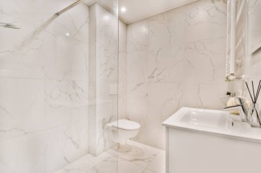 Beyaz mermer duvarları ve zemini olan bir banyo. Duş kabininin yanındaki tuvalet de dahil.