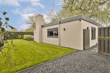 Mavi bir gökyüzünde, arkasında ağaçlar ve çalılar olan yeşil bir çimenliğin ortasında küçük bir ev.