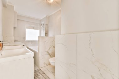 Duvarlarında mermer döşeme ve beyaz eşyalar olan bir banyo. Köşesinde de bir küvet.