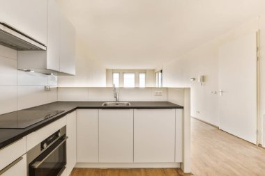 Adanın önünde beyaz dolapları ve siyah tezgahları olan boş bir mutfak da görülebilir.