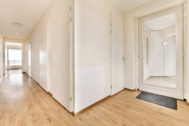 Ahşap zeminleri ve beyaz duvarları olan boş bir oda. Kapının önündeki duvarda büyük bir ayna var.