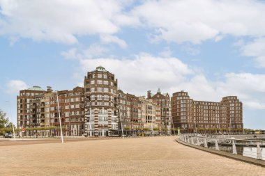 Su kenarında mavi gökyüzü ve beyaz bulutlu eski bir tuğla bina çekilmiştir.