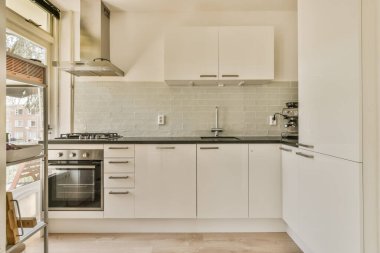 Mutfak, beyaz dolaplar ve aletler tezgahın üstünde fırının önünde, lavabo ve bulaşık makinesi.