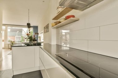 Görüntünün ortasında beyaz dolapları ve siyah tezgahları olan modern bir mutfak ocağın üstünde bir fırın kaputu bulunur.