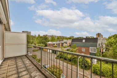 Arka planda ağaçlar ve binalar olan bir balkon. Bir apartmanın penceresinden sokağa bakıyor.