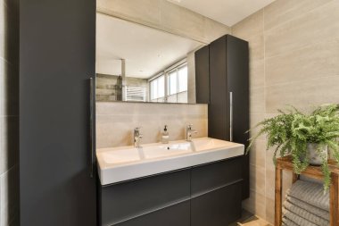 Modern bir banyo, siyah dolaplar ve beyaz tezgahlar, lavaboda bir bitkiyle birlikte aynanın yanında.