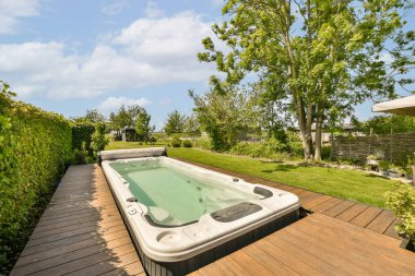 Bahçedeki ahşap bir güvertede yeşil ağaçlar ve çalılarla çevrili bir jakuzi berrak mavi gökyüzü.