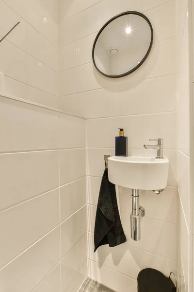 a bathroom with white tiles and black towel hanging on the wall next to a sink bowl, mirror and toilet brush