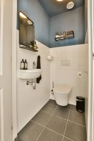 シンクの白いトイレを含む青い壁とタイルのフロアーリングが付いている小さなバスルームは鏡の隣にあります — ストック写真