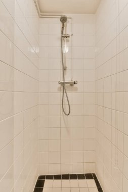 Beyaz fayanslı banyo, yerde siyah beyaz fayanslar, duş başlığı ve köşede el parmaklığı.