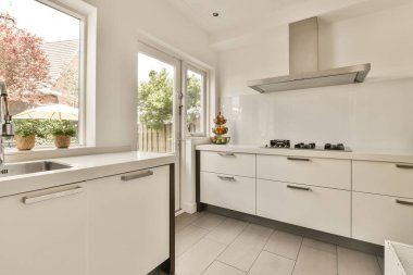 Tezgahın üstünde beyaz dolaplar ve aletler olan bir mutfak. Dışarıdaki verandaya bakan büyük bir pencerenin önünde.