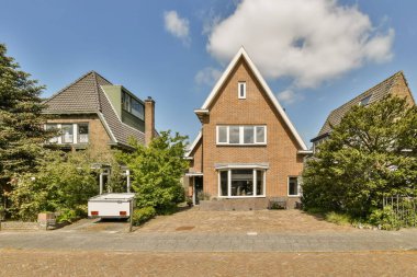 Her iki tarafında ağaçlar ve çalılar olan bir alanın ortasında bir tuğla ev, mavi bir gökyüzü var.