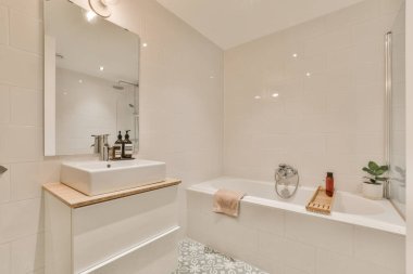 Duvarlarında beyaz fayanslar ve döşemeleri olan bir banyo. Küvetin üstünde büyük bir ayna var.