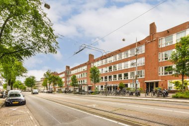 Kenarında arabaları olan bir şehir caddesi ve apartmanların önünde yürüyen insanlar.